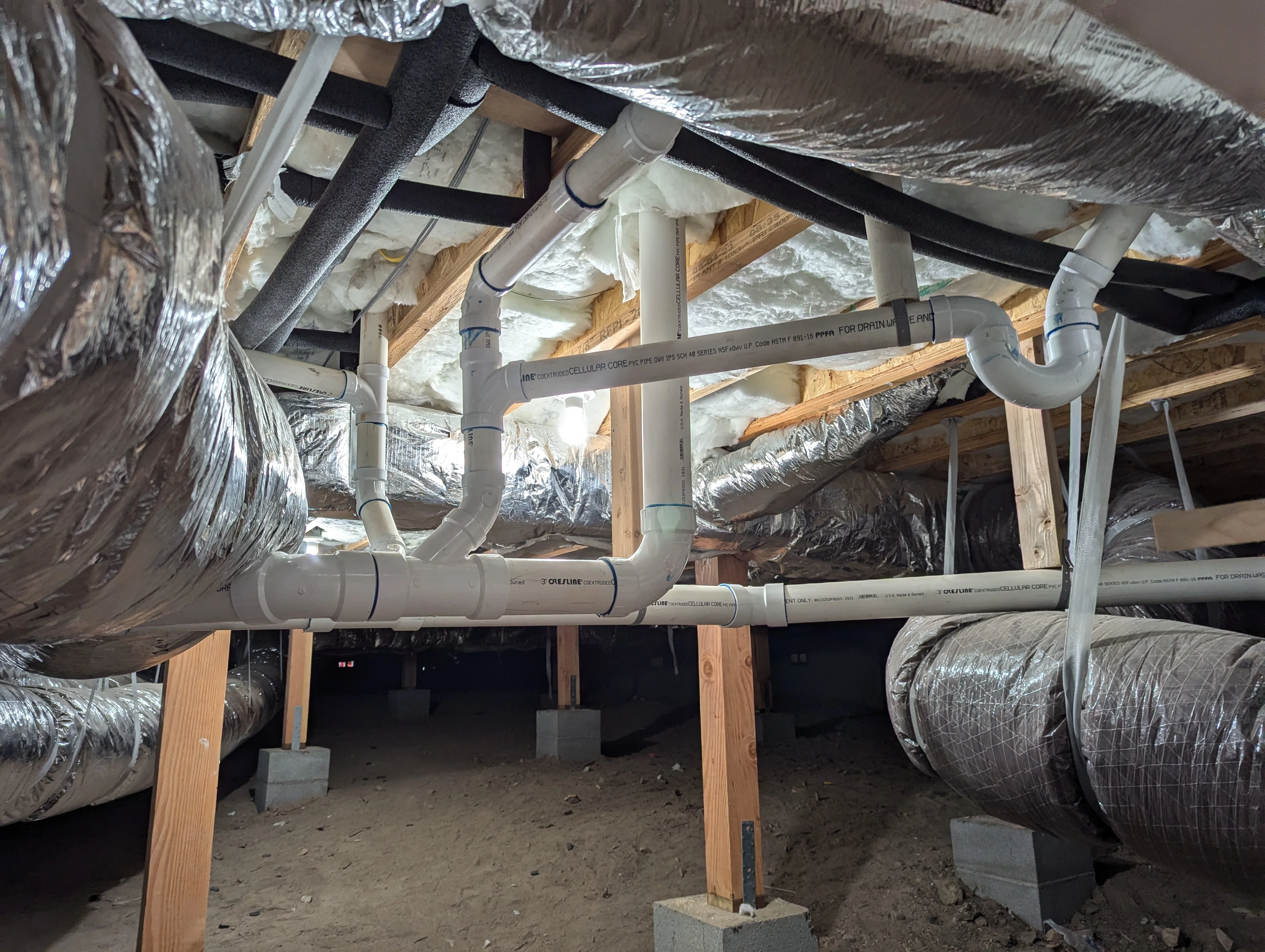 PVC drain and vent pipes installed in crawlspace with wooden framing and insulation wrap